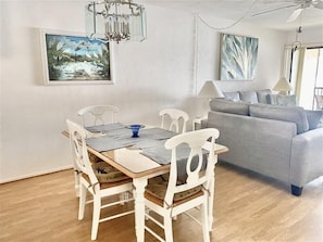 A dining area with a wooden table and white chairs, a grey sofa, and two paintings on the walls. - Runaway Bay 239-Bayfront view - Pool, tennis, pickleball-Resort style community (Bradenton Beach)