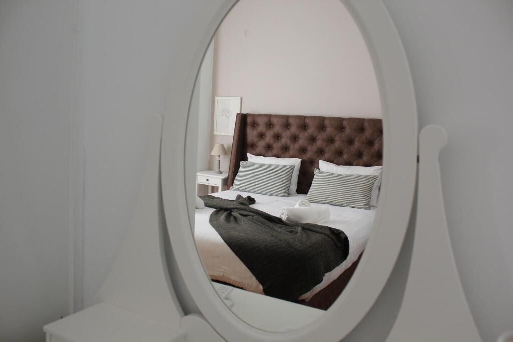 A bedroom with a tufted headboard, a bed with striped and solid pillows, a nightstand with a lamp, and a window with a view of the room.