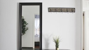 Een spiegel met een zwarte lijst, een klein potplantje op een zwarte poef, en een decoratieve wanddecoratie met haken.
