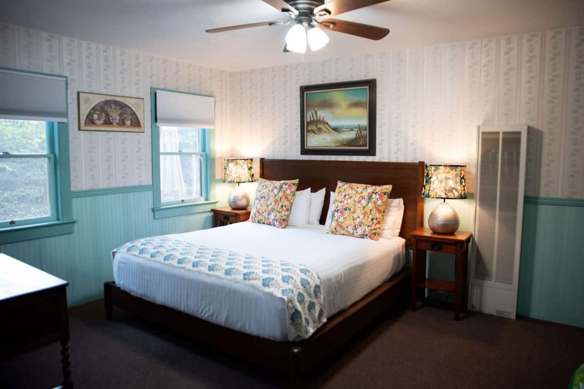 A bedroom with a large bed, two bedside tables with lamps, a ceiling fan, a window with blinds, and a framed picture on the wall.