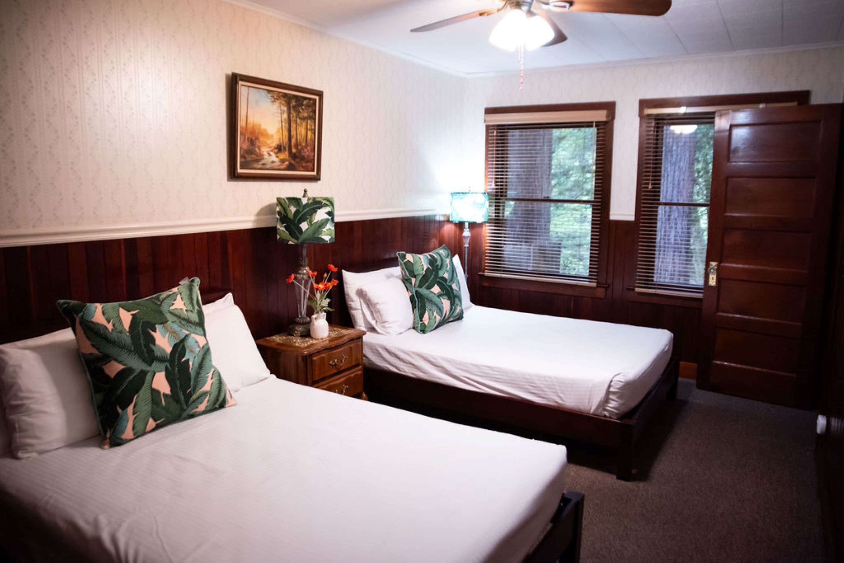 A room with two beds, a ceiling fan, a painting on the wall, and a window with blinds.