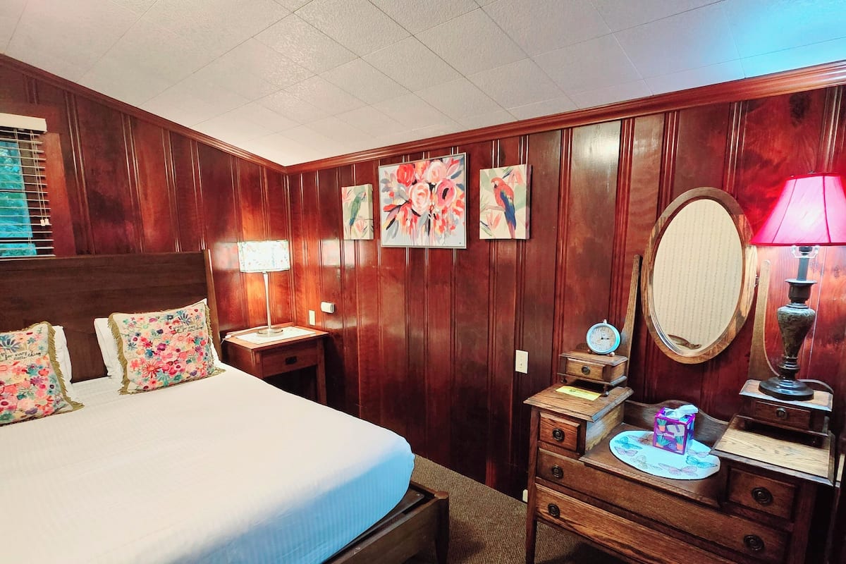 A hotel room with a large bed, wooden paneling, a dresser with a mirror, and floral artwork on the walls.