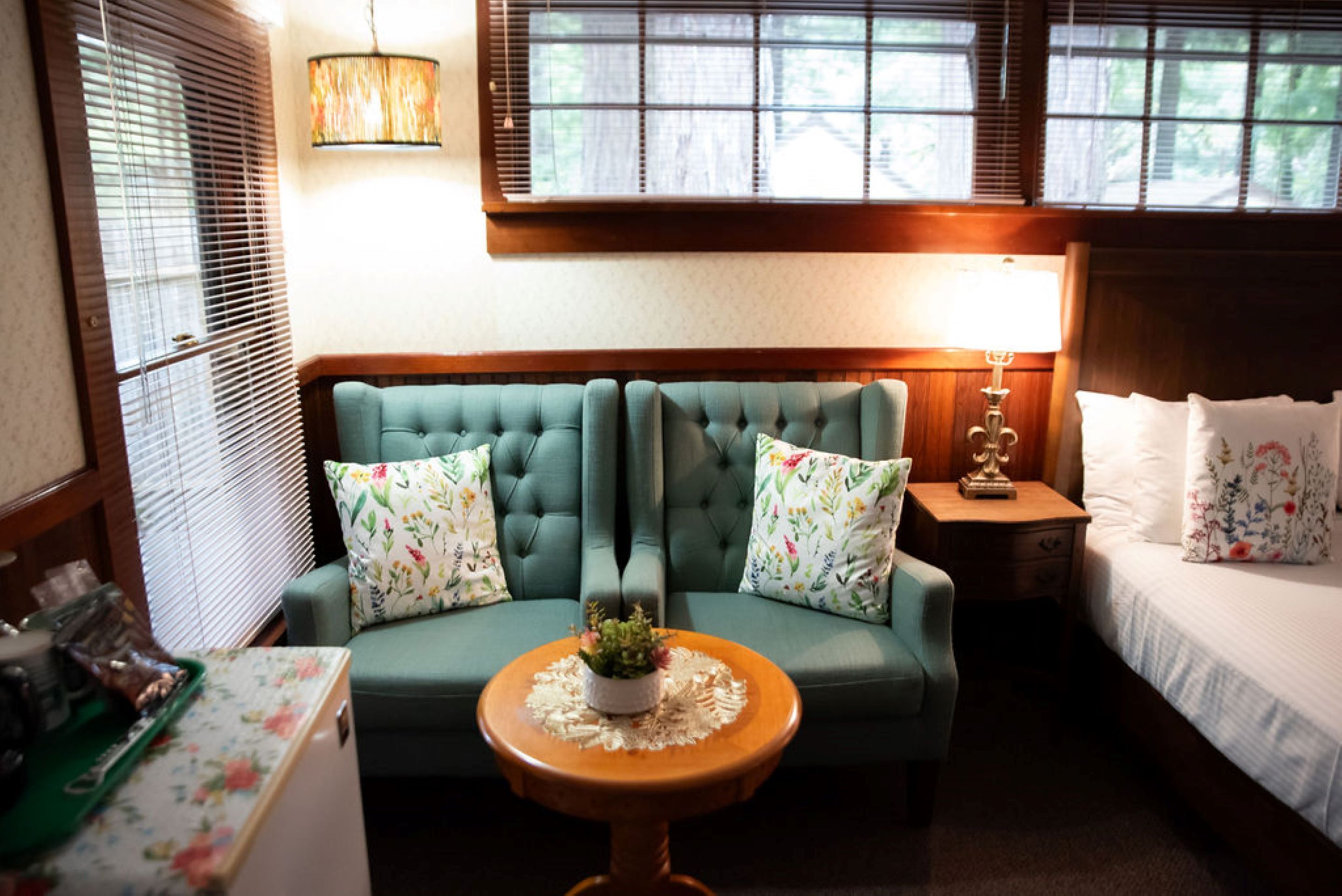 A cozy room with a green sofa, a wooden nightstand, a small table with a plant, and a bed with floral pillows.