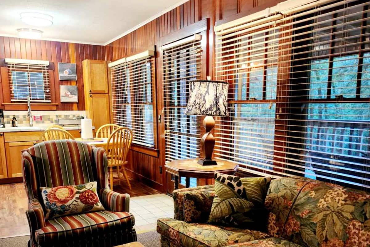 A cozy living room with a striped armchair, a floral sofa, and a stone coffee table. There is a kitchen in the background with wooden cabinets and a window with blinds.