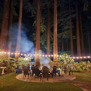 An outdoor seating area with chairs arranged around a table, surrounded by tall trees and string lights. - Miranda Gardens Resort (Myers Flat)