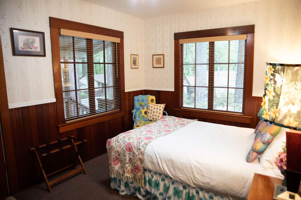 A bedroom with two windows, a bed with floral bedding, a chair, and a small table.