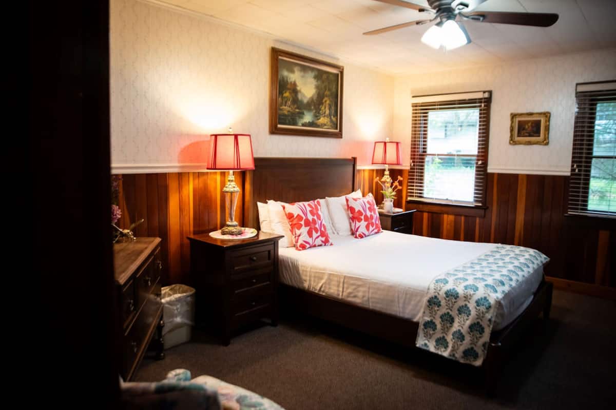 A bedroom with a bed, two bedside tables with lamps, a ceiling fan, a window with blinds, and a framed picture on the wall.