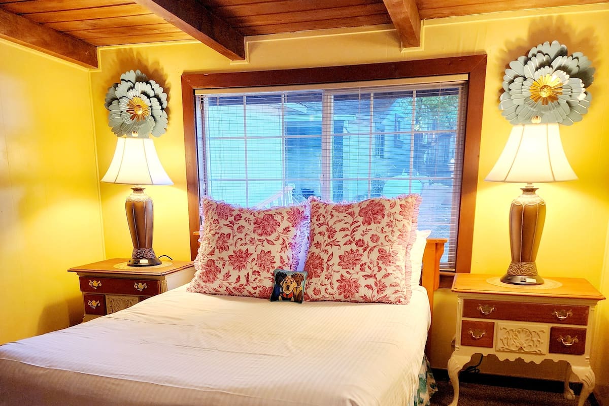 A neatly made bed with floral bedding, two bedside lamps, and a window with blinds.