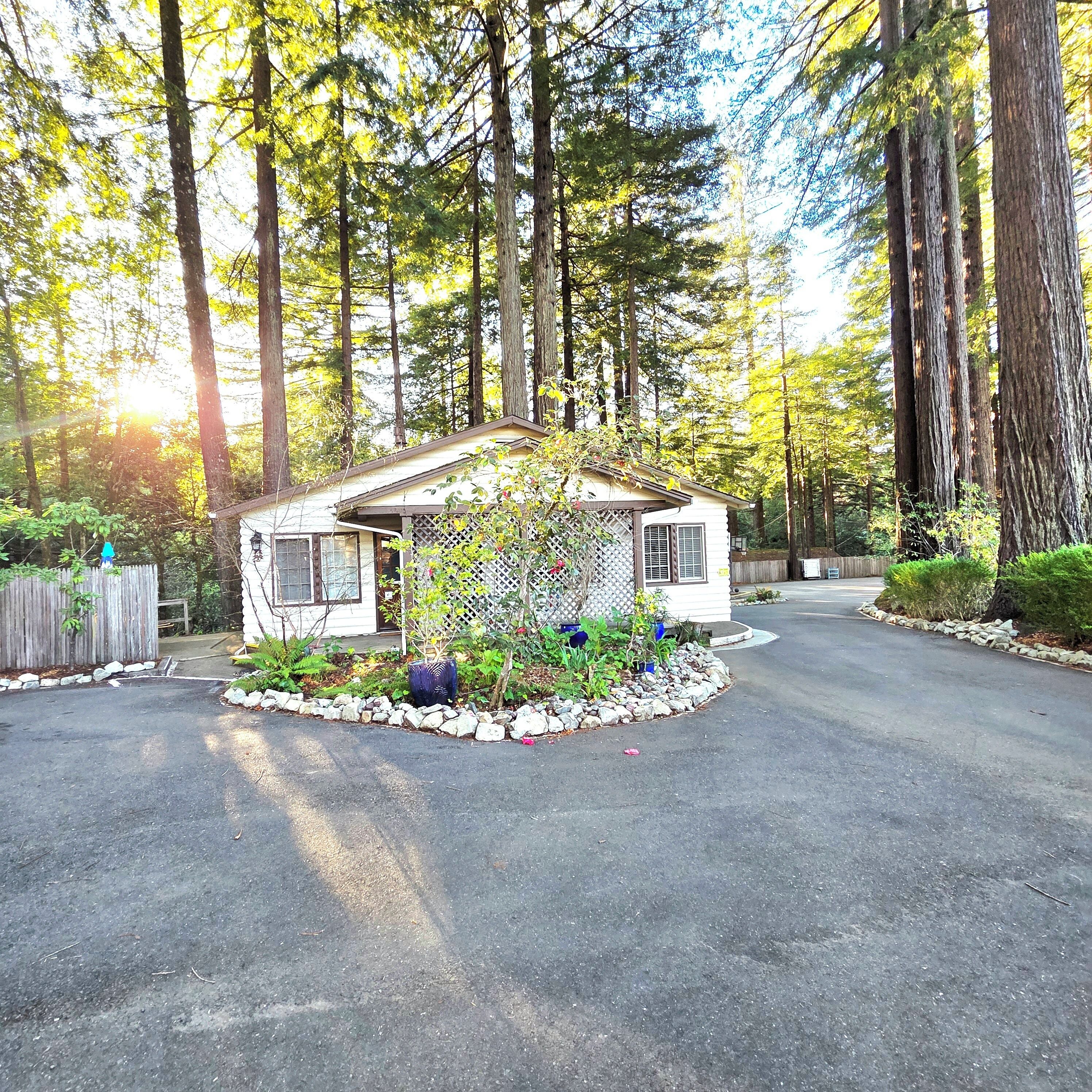 A small house with a porch, surrounded by a stone border, in a forested area.