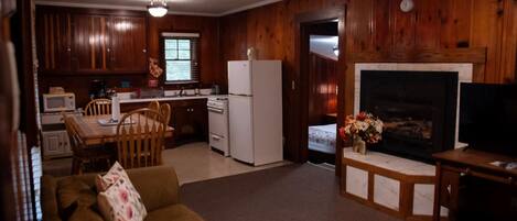 A cozy living room with a fireplace, wooden walls, a dining area with a table and chairs, and a kitchen with a refrigerator and microwave.