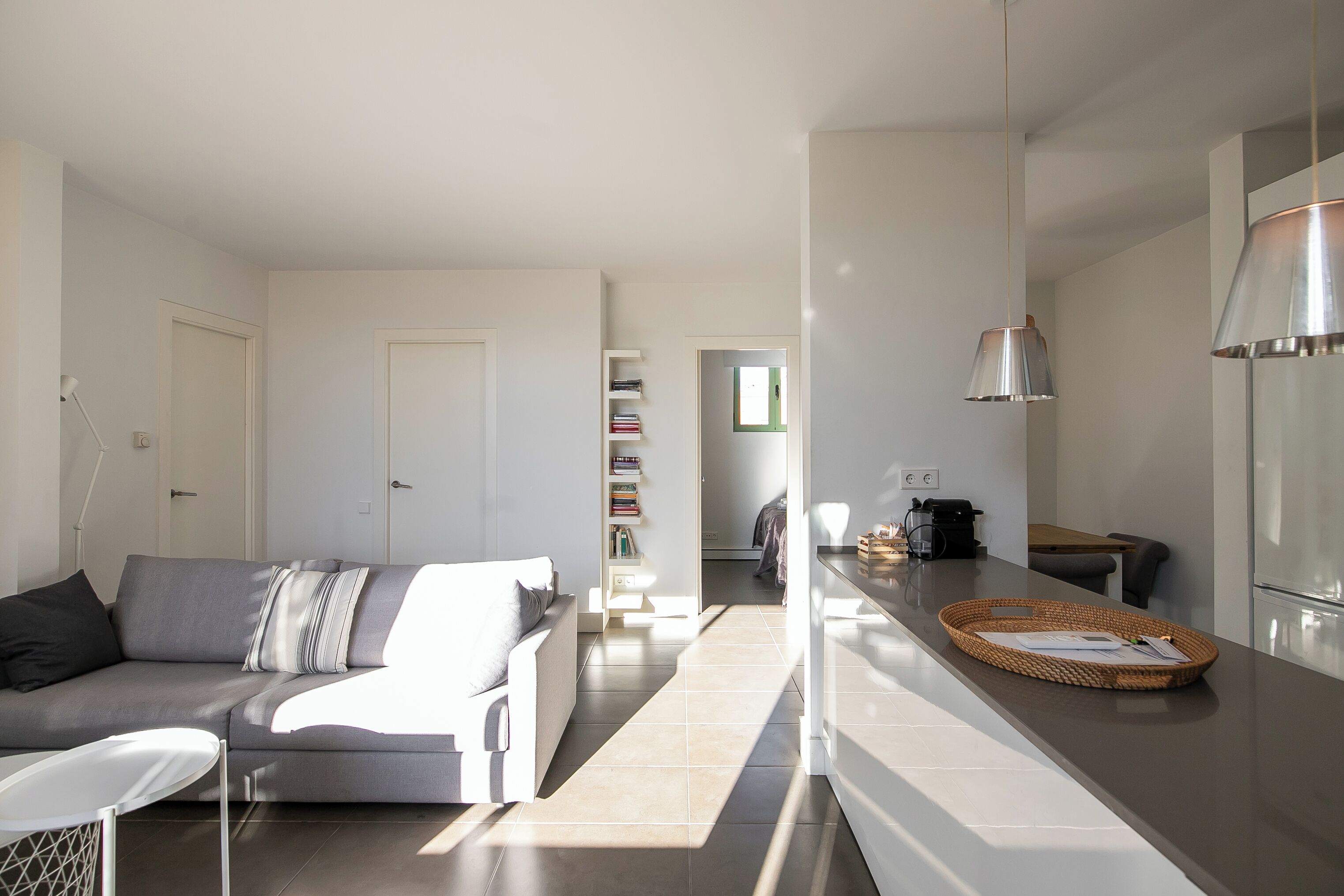 Una sala moderna con un sofá gris, un sillón blanco y una cocina con una encimera y una lámpara colgante.