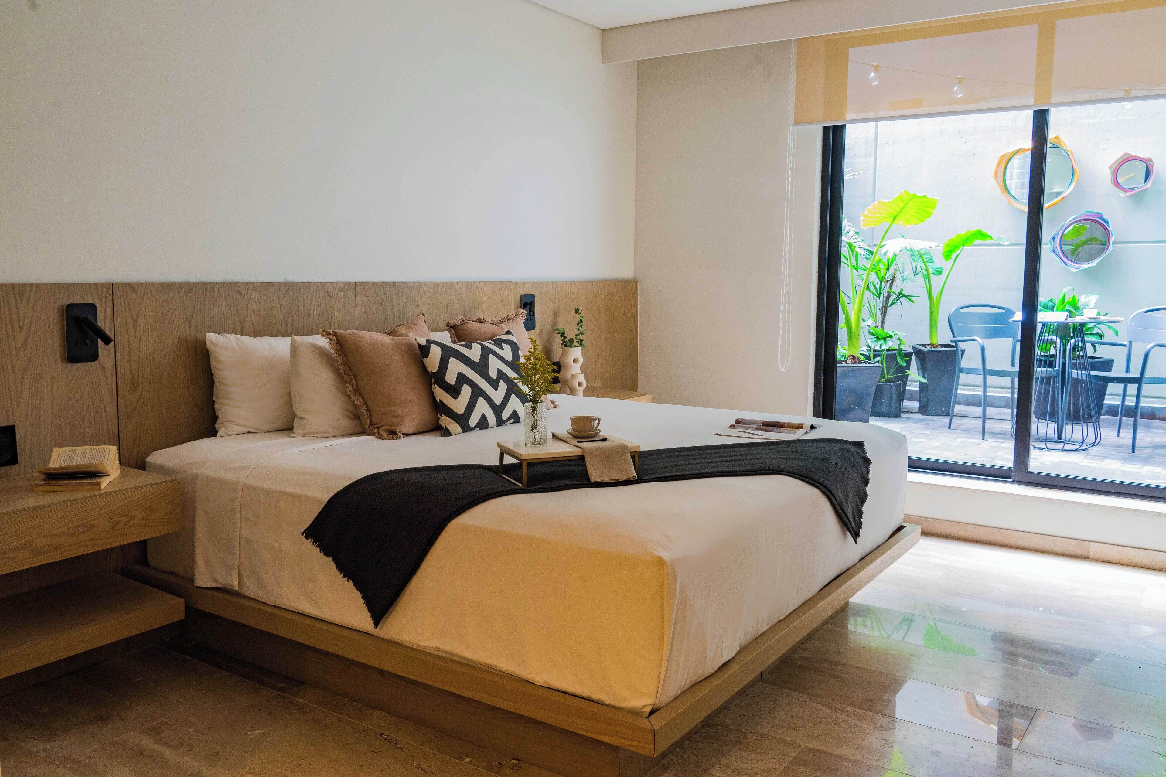 A modern bedroom with a bed, bedside table, and a view of the outdoors through large windows.