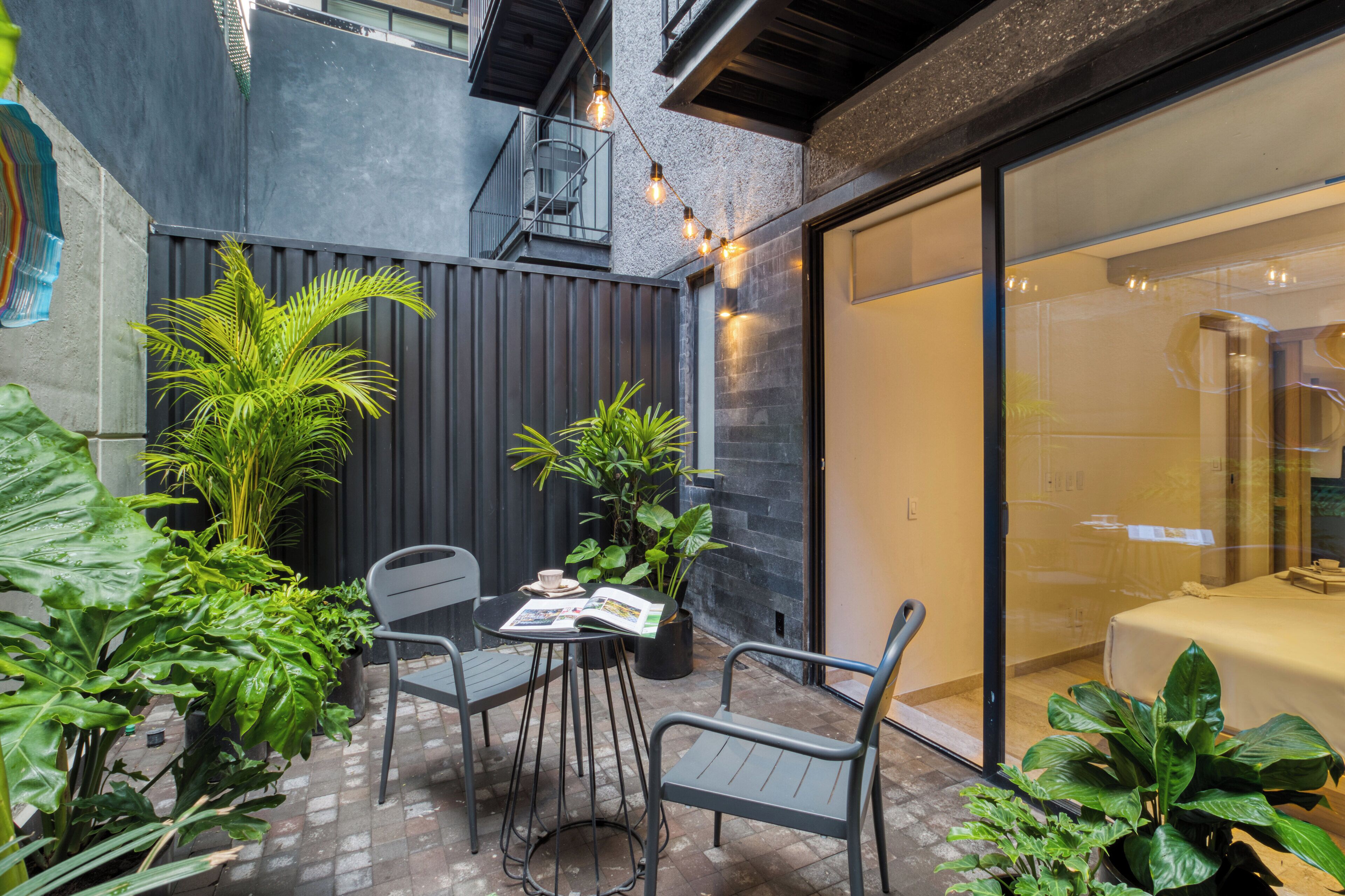 En lille patio med borde og stole, omgivet af plantekasser og en moderne bygning med en glasdør.