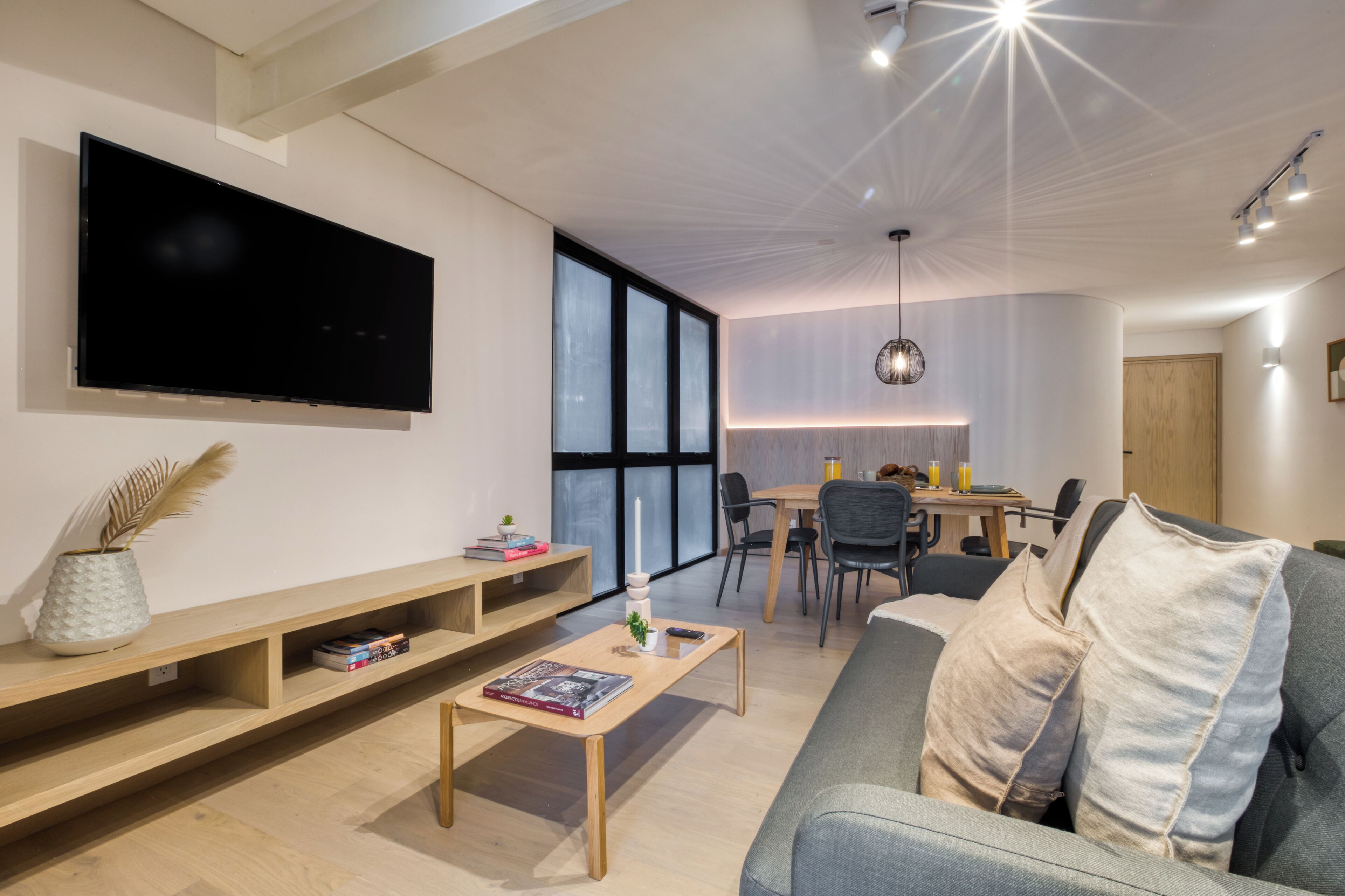 A modern living room with a flat-screen TV, wooden furniture, a dining area, and large windows.