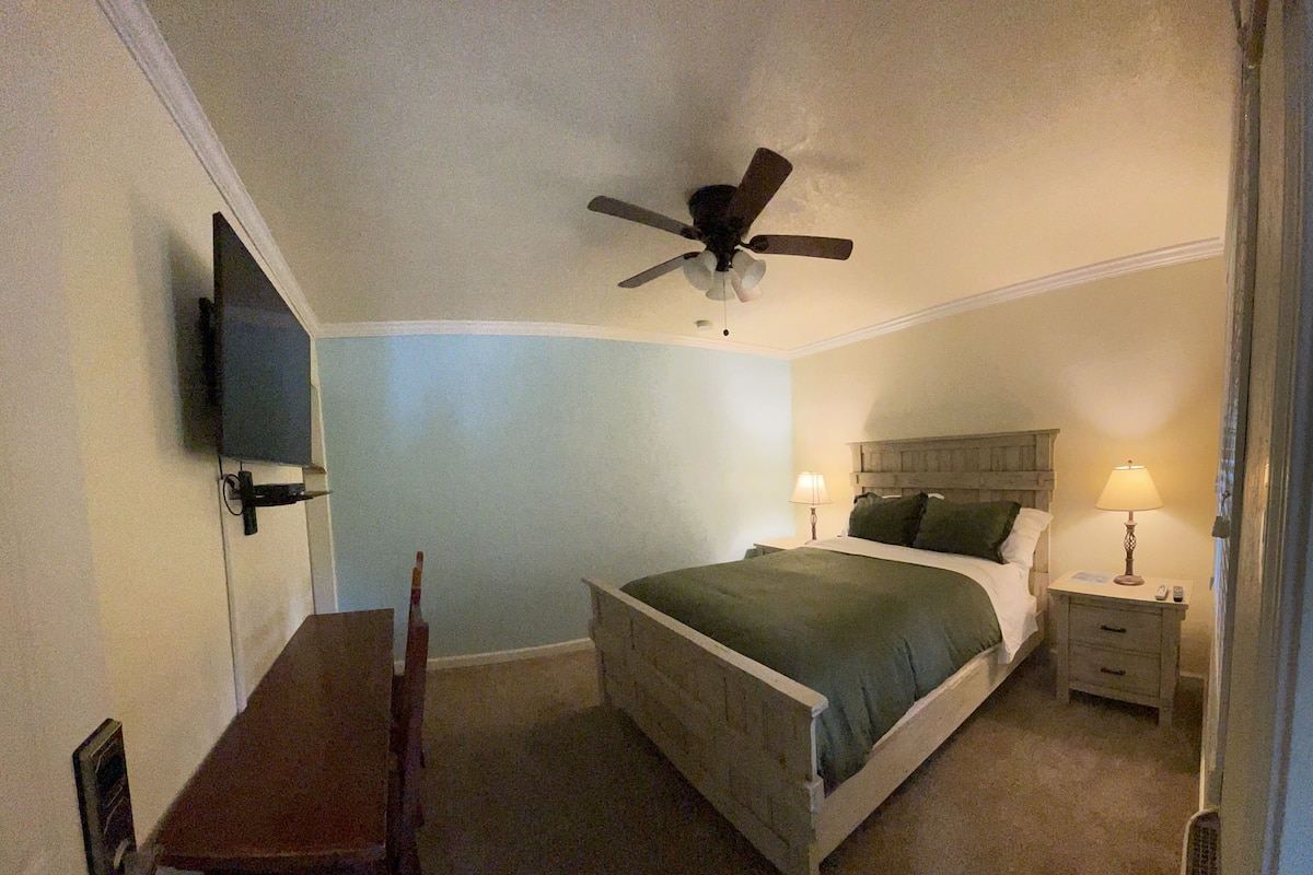 A bedroom with a bed, bedside tables, a desk, a chair, a ceiling fan, and a TV mounted on the wall.