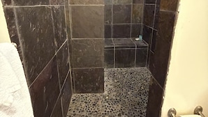 A bathroom with dark stone tiles, a shower area with a pebble floor, and a toilet with a paper towel holder.