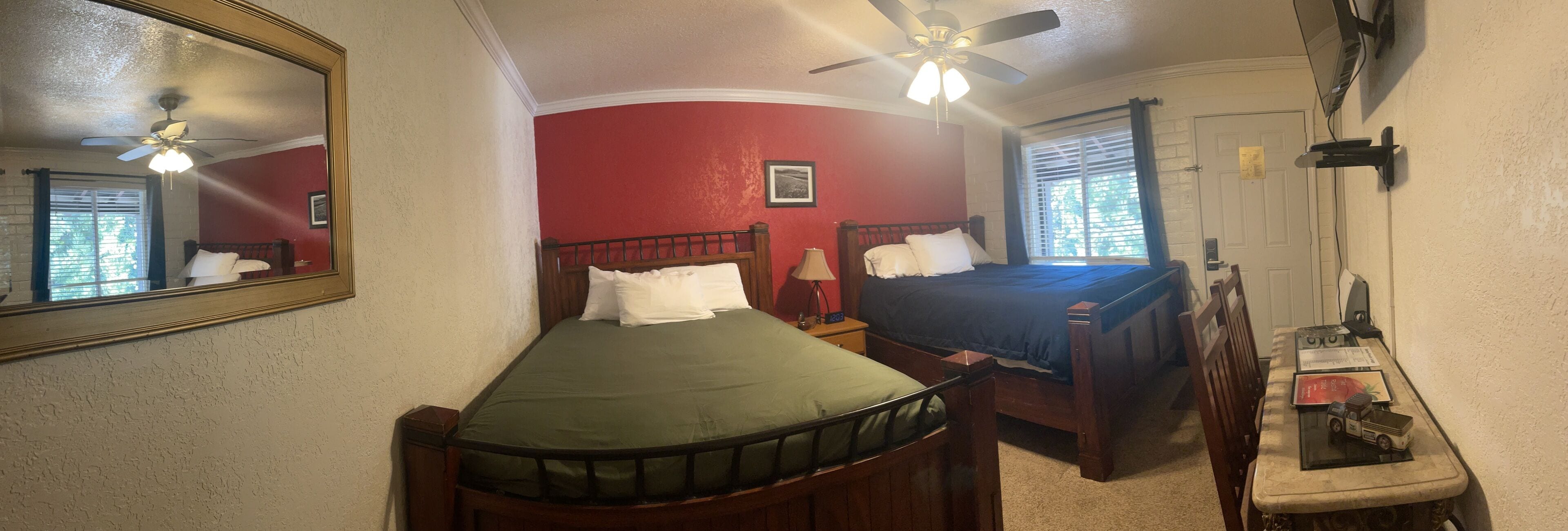 A room with two beds, a ceiling fan, a desk with a lamp, and a window with blinds.