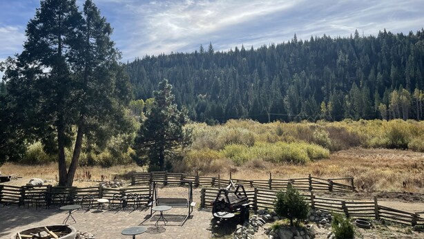 An outdoor seating area with tables and benches, surrounded by a wooden fence, overlooking a forested hillside.