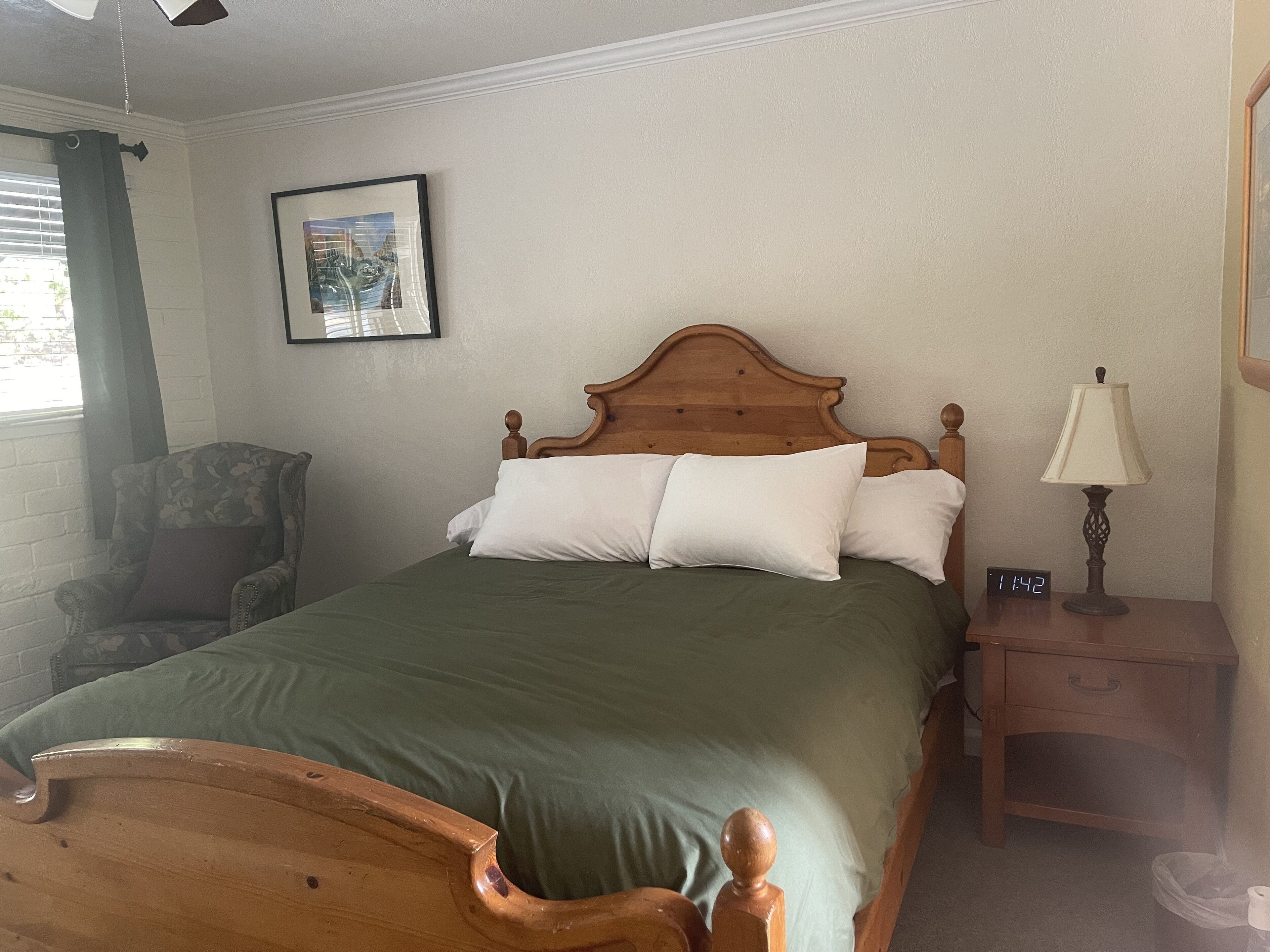 Een net opgemaakt bed met een houten hoofdbord, een groene dekbedovertrek en witte kussens. Links van het bed staat een stoel met een bloemenprint. Daarboven hangt een ingelijst schilderij aan de muur.