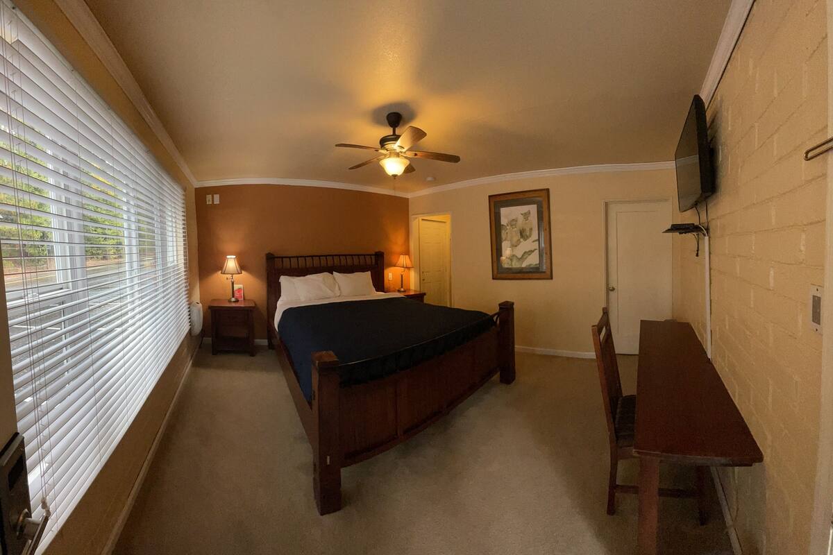A bedroom with a bed, a desk, a chair, a ceiling fan, and a window with blinds.