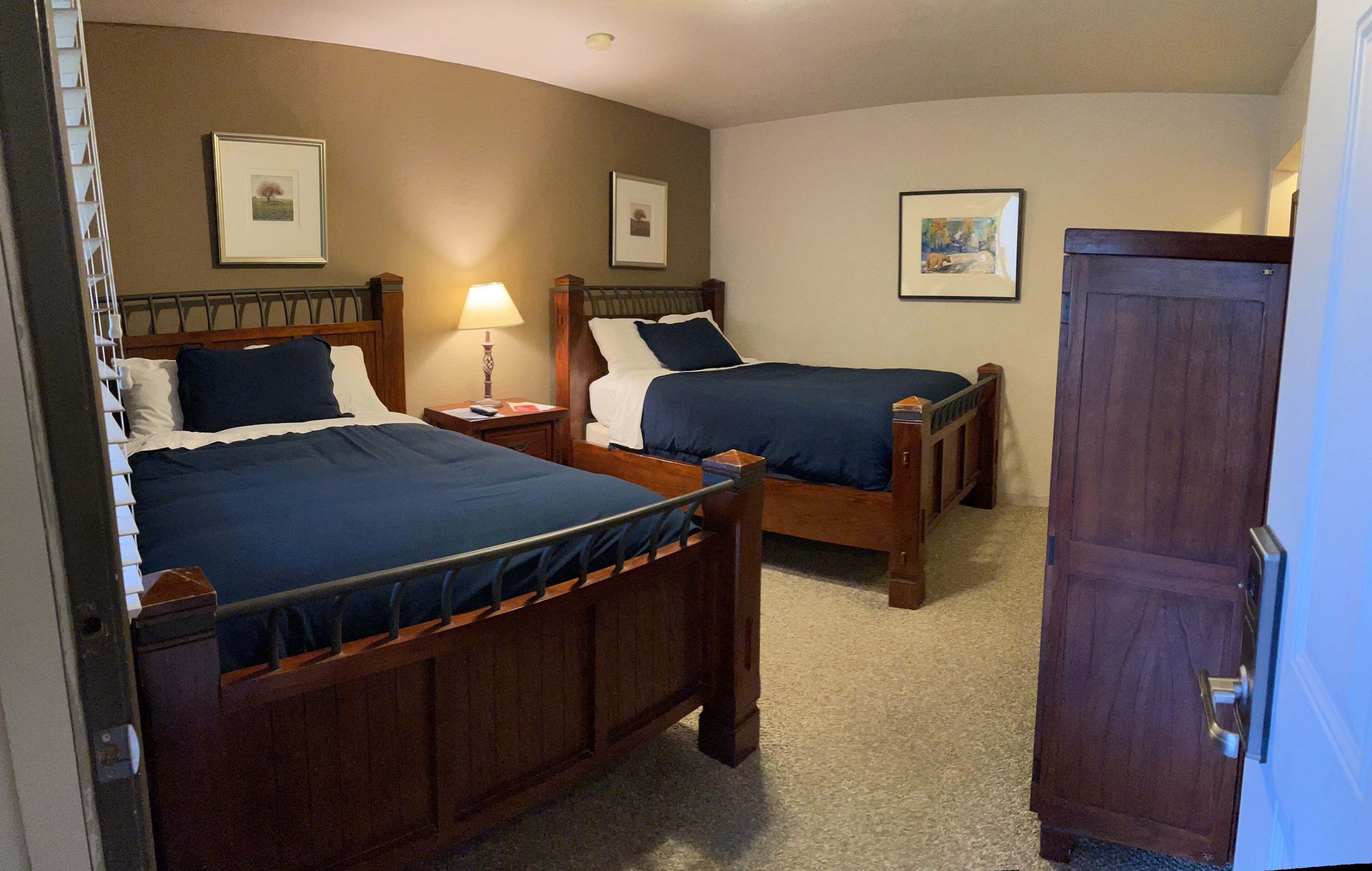 A hotel room with two wooden beds, a nightstand with a lamp, and a wardrobe.