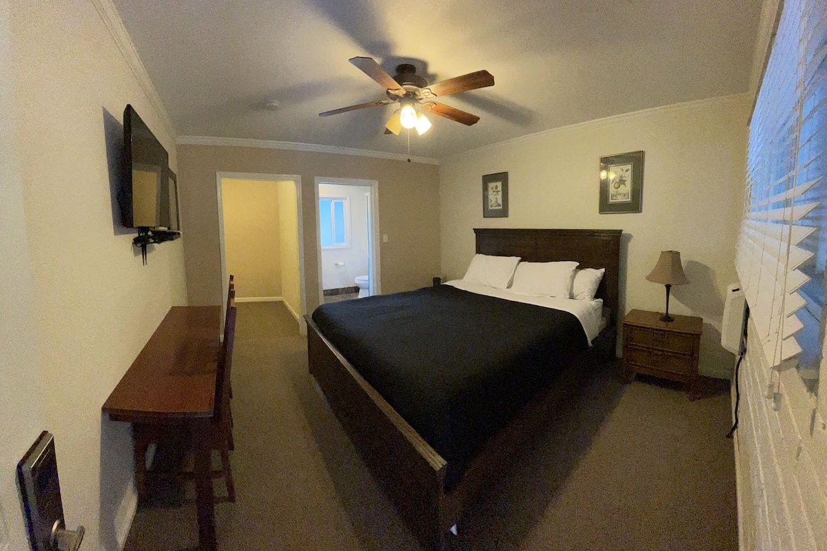 A bedroom with a bed, a desk, a chair, a nightstand, a lamp, a ceiling fan, and a TV on the wall.