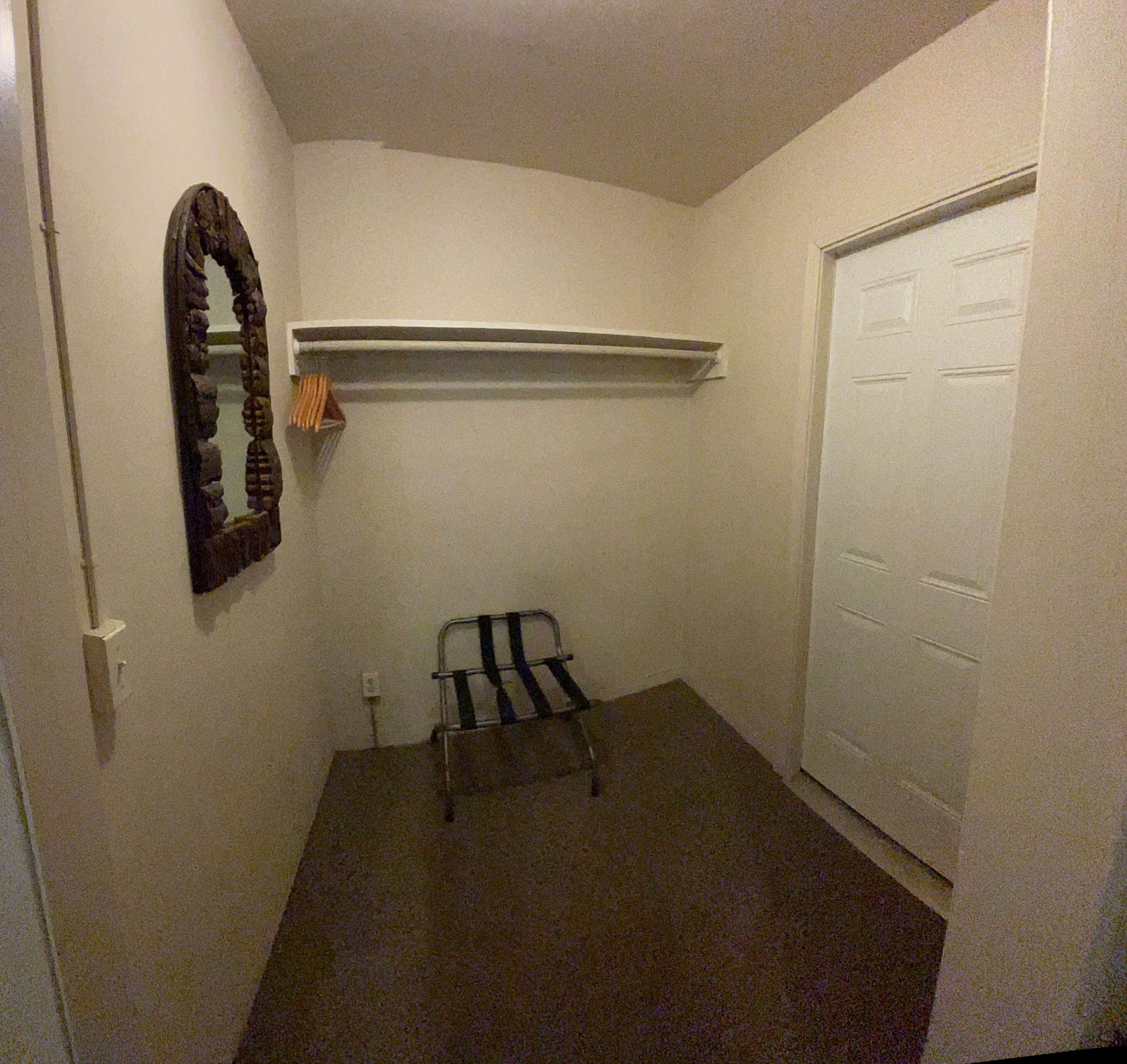 Um quarto pequeno e pouco iluminado, com porta, espelho, prateleira e uma cadeira.