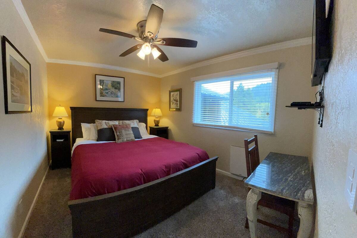 A bedroom with a bed, two bedside tables, a ceiling fan, a window with blinds, and a small table with a chair.