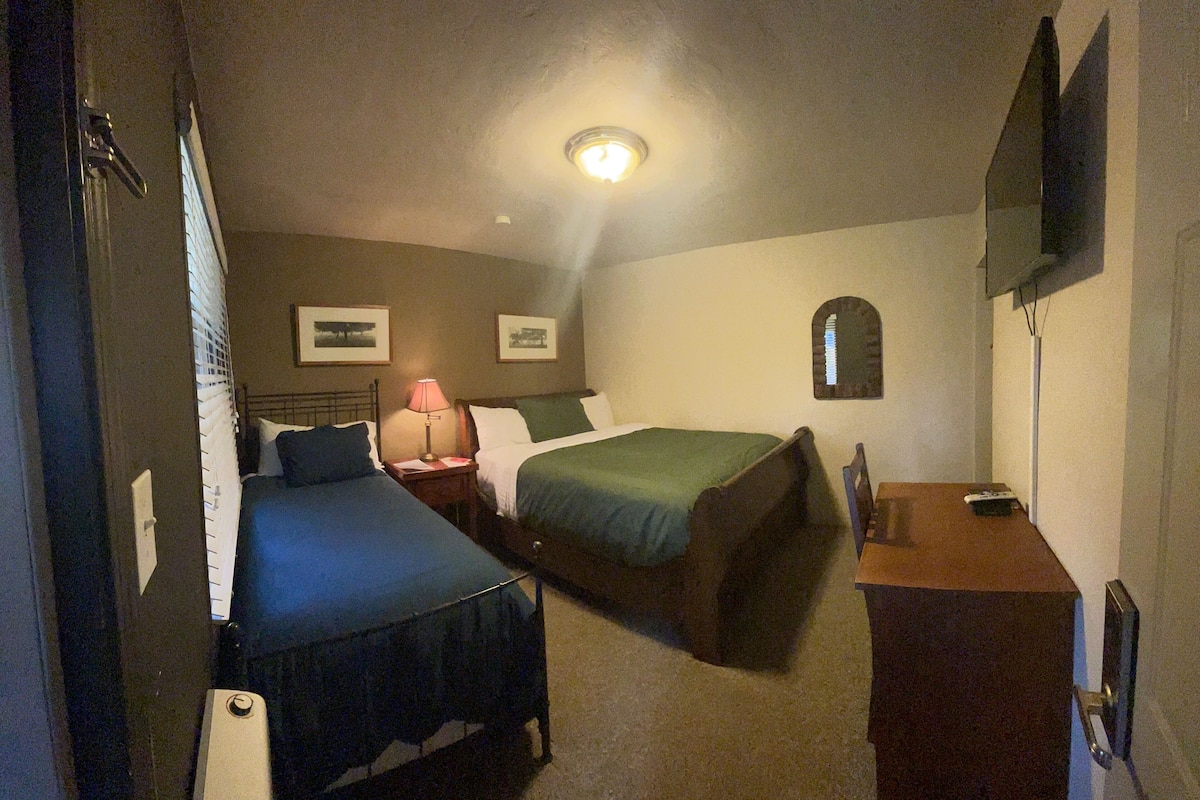 A hotel room with two single beds, a desk, a chair, a lamp, and a television mounted on the wall.