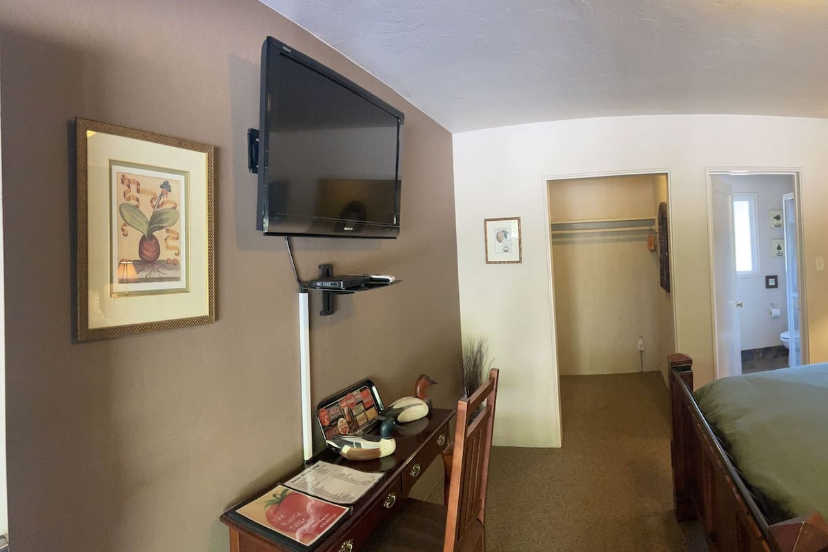 A hotel room with a bed, a desk, a chair, a TV, a window with blinds, and a door leading to another room.