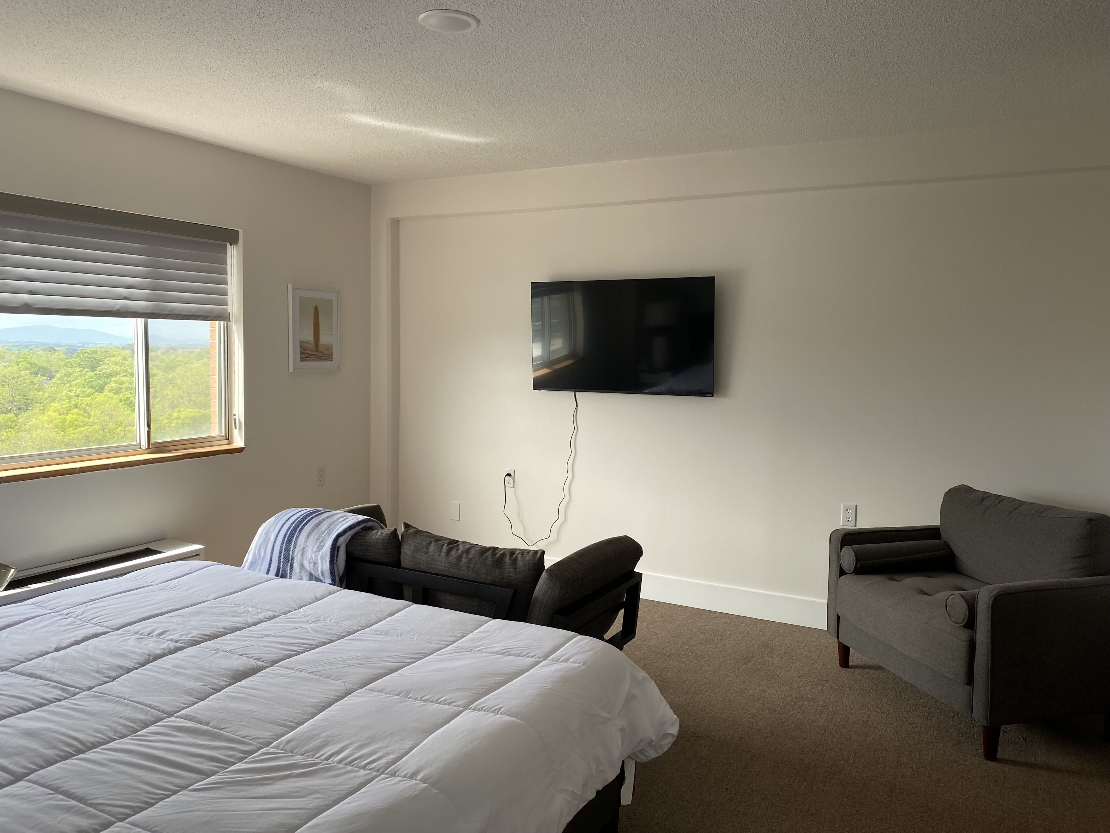 Une chambre d’hôtel avec un lit, une chaise, un téléviseur fixé au mur et une fenêtre avec des stores.