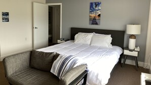 Une chambre d’hôtel bien rangée, avec un lit, une chaise, une table de chevet avec une lampe et un tableau au mur.