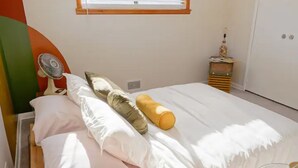 A neatly made bed with white and pink bedding, a yellow pillow, and a white pillow. There is a small bedside table with a lamp and a decorative item. A window with blinds is visible in the background. - Retro home minutes from downtown Denver (Denver)