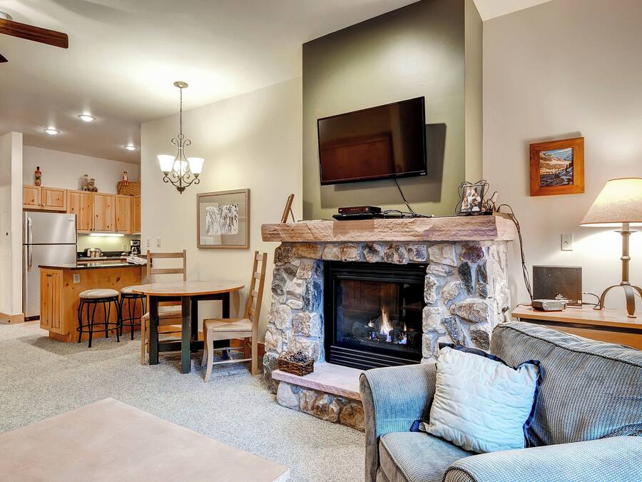 Una sala de estar acogedora con chimenea de piedra, televisor montado en la pared y comedor con alacenas de madera y refrigerador.