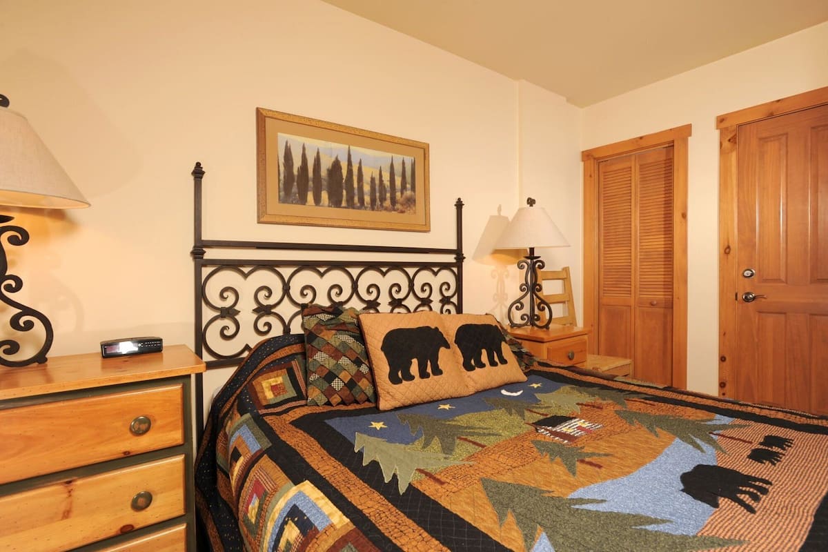 A bedroom with a bed, a nightstand, a dresser, a lamp, a framed picture, and a wooden door.