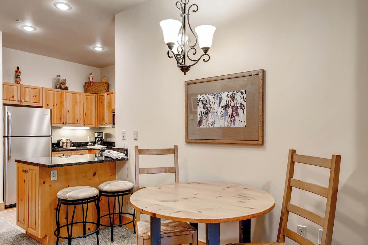 A kitchen with wooden cabinets, a stainless steel refrigerator, a round wooden table with two barstools, and a chandelier.