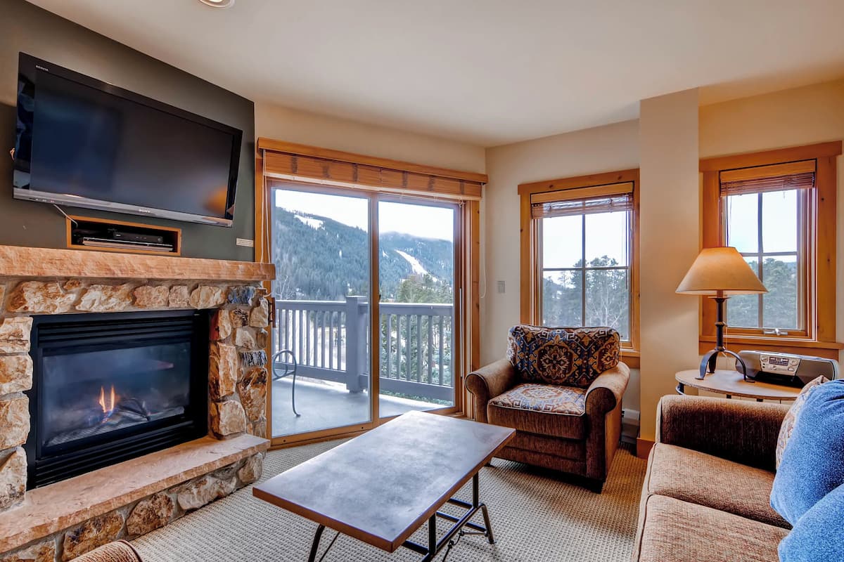 A living room with a stone fireplace, a flat-screen TV, a sofa, a coffee table, and a balcony with a view of the mountains.