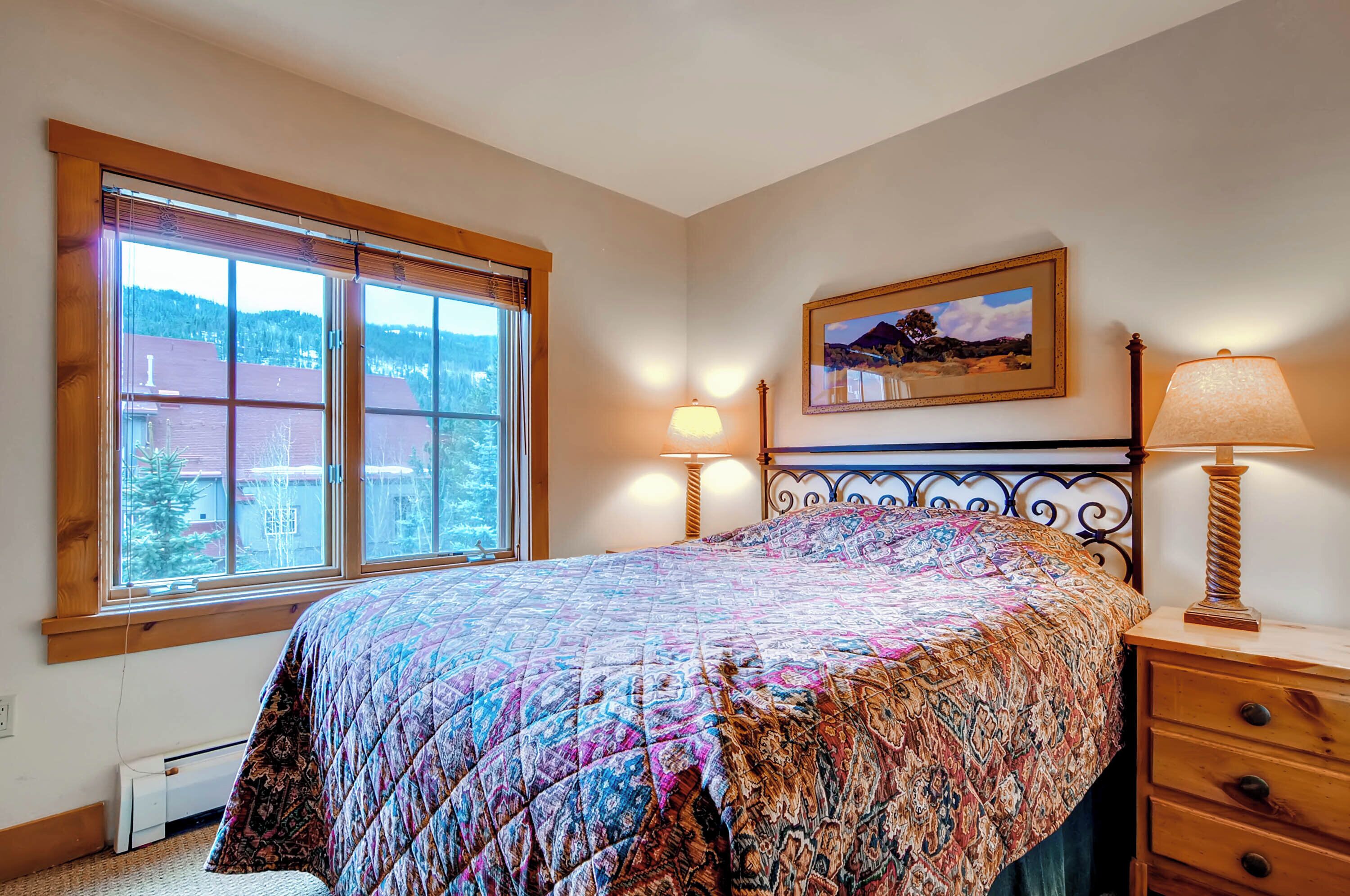 A bedroom with a large window, a bed with a patterned quilt, two bedside lamps, a wooden nightstand, and a framed picture on the wall.