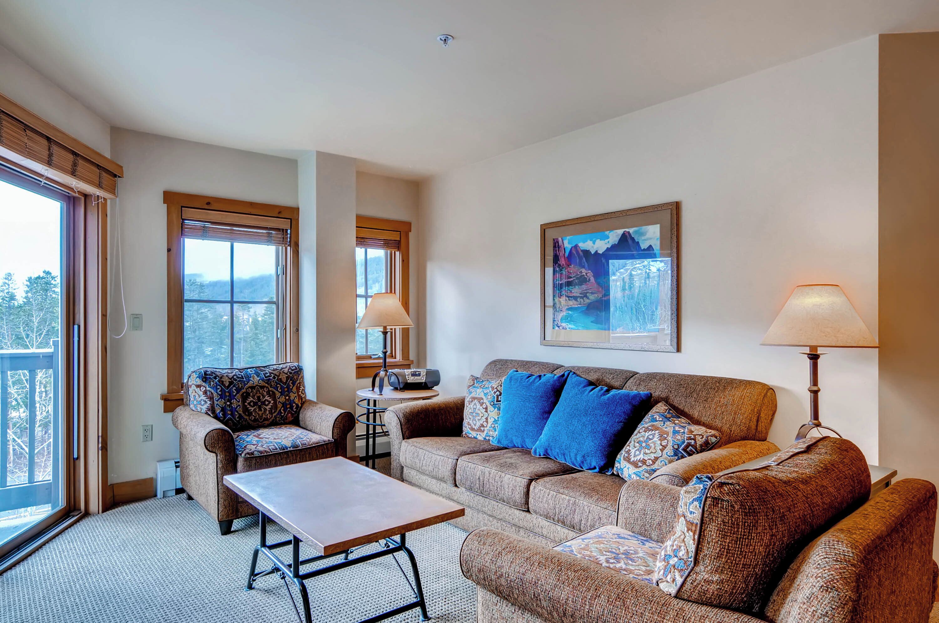 A cozy living room with a sofa, armchair, and coffee table. There are two windows with blinds, a lamp, and a framed picture on the wall.