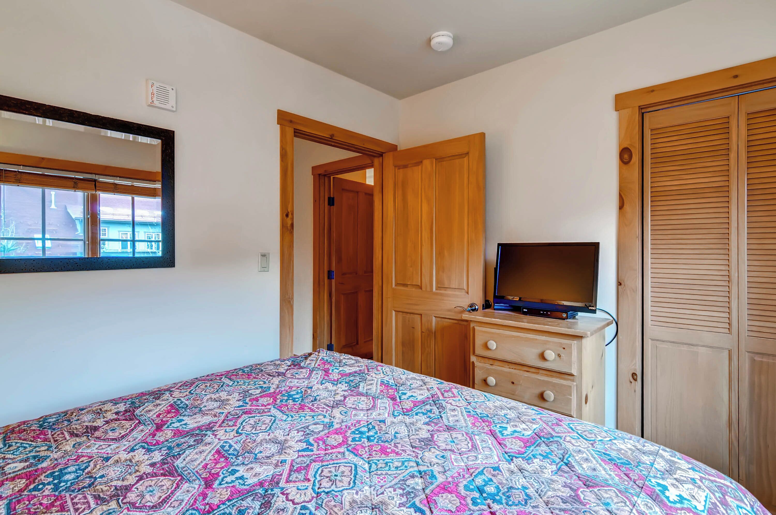 Een slaapkamer met een bed met een decoratieve spreideken, een houten dressoir met een televisie, een spiegel en een houten deur.