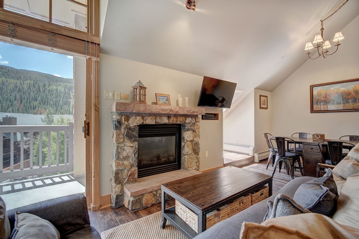 A spacious living room with a stone fireplace, a dining area, and a balcony with a view of a forested mountain.