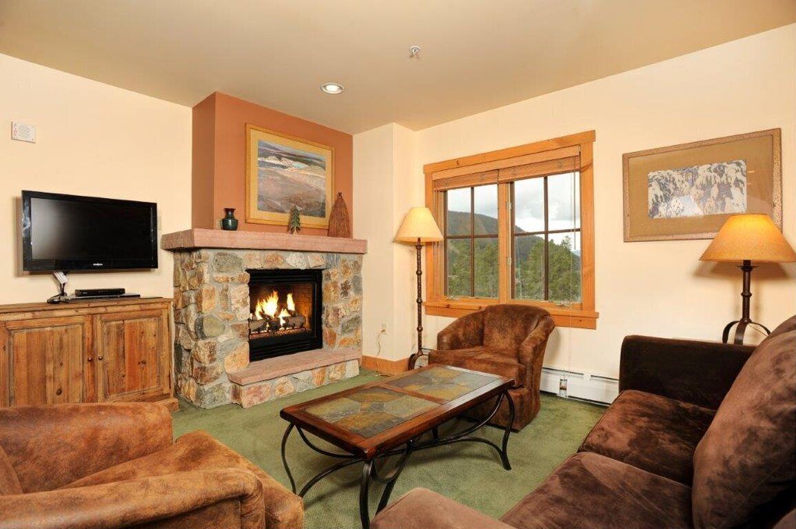 A cozy living room with a stone fireplace, leather sofas, a flat-screen TV, and a large window with a mountain view.