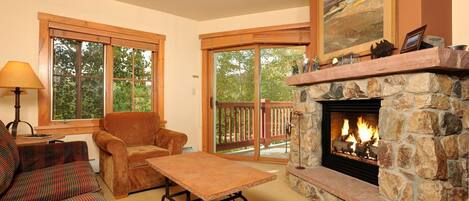 A cozy living room with a stone fireplace, a sofa, a coffee table, and large windows offering a view of the outdoors.