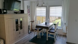 A dining area with a table and chairs, a television on a cabinet, and a view of the outdoors through large windows. - Charmantes 60m² Ferienhaus, Ausgestattet mit Terrasse (Zinnowitz)