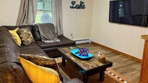 Ein Wohnzimmer mit braunem Ledersofa, einem Holztisch, einem an der Wand befestigten Fernseher und einem Fenster mit Vorhängen.