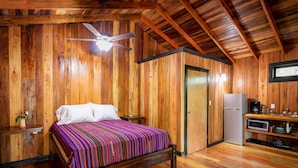 Une pièce aux murs lambrés, comprenant un lit, un ventilateur de plafond, un réfrigérateur et une petite kitchenette.