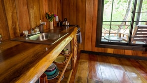 Un intérieur en bois comprenant un coin cuisine avec un évier, une fenêtre donnant sur un espace extérieur aménagé, et une étagère avec de la vaisselle.