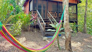 Une cabane en bois dotée d’une terrasse en bois et de grandes fenêtres, entourée d’une végétation luxuriante et agrémentée d’un hamac.