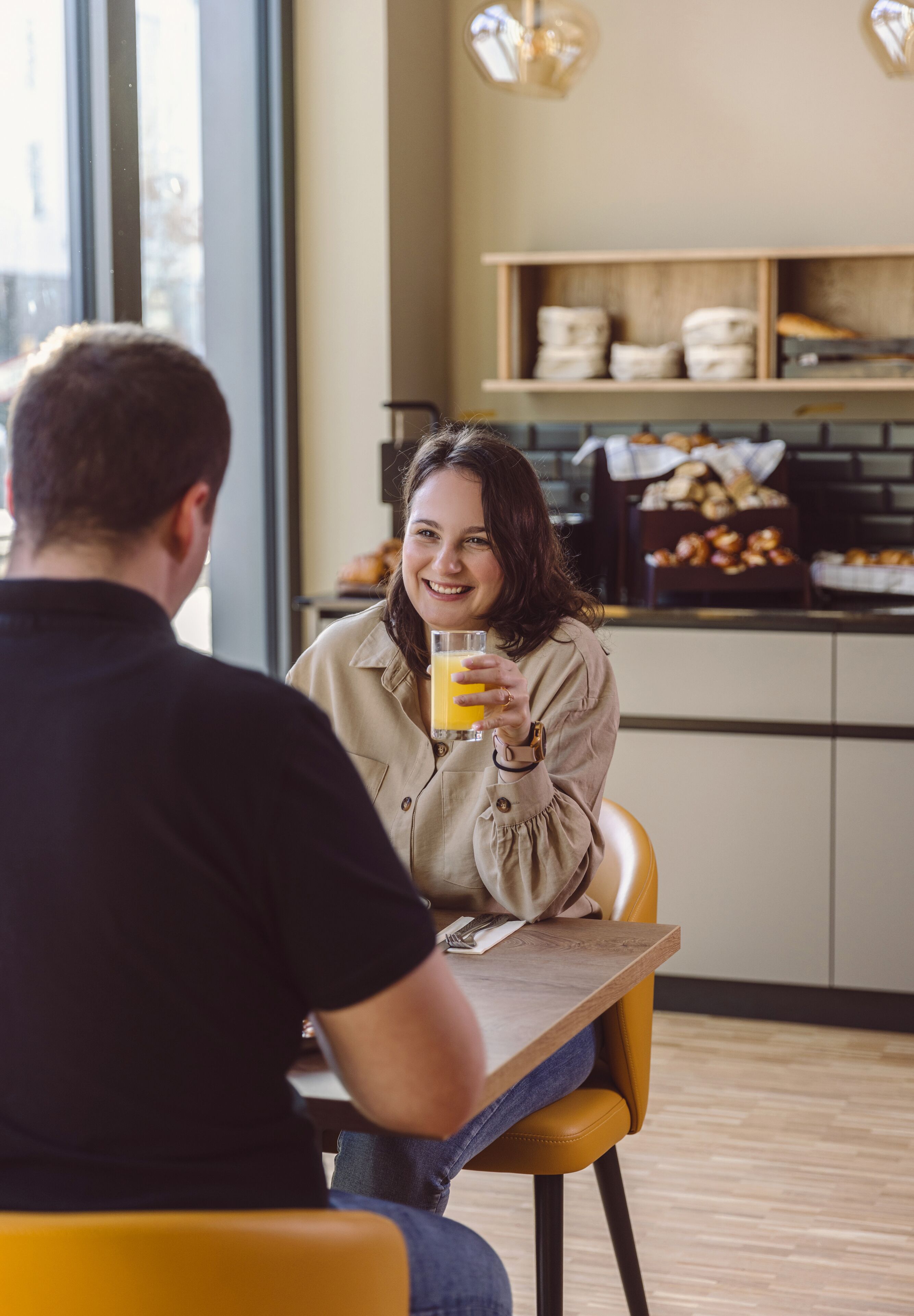 Un coin repas avec une table et des chaises, une personne tenant un verre de jus d’orange, et un espace cuisine en arrière-plan.