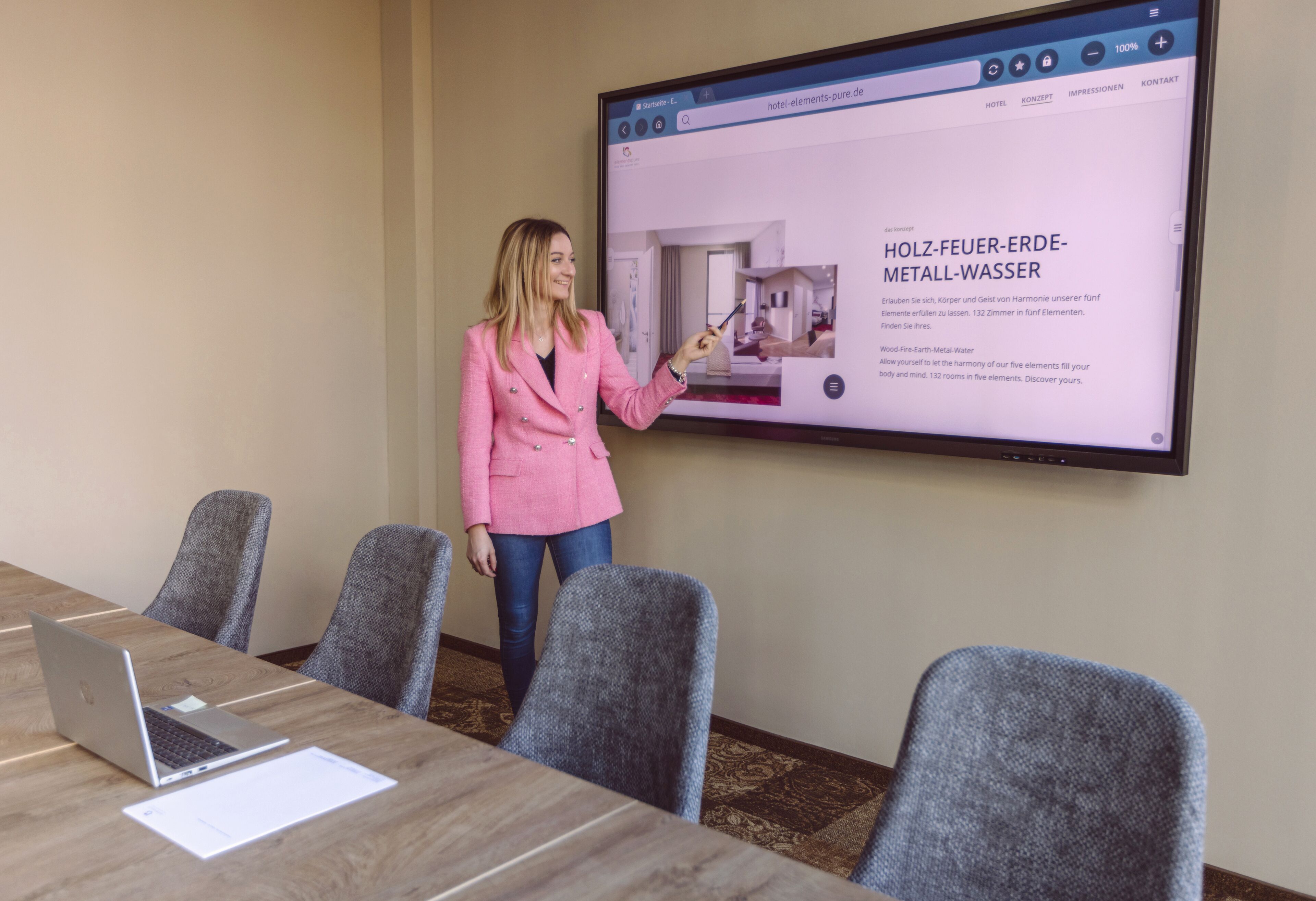 A presentation in a conference room with a laptop and documents on the table, a large screen displaying hotel information, and a person presenting.